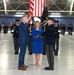 A formal military swearing-in or commissioning ceremony taking place inside what appears to be a large aircraft hangar. A civilian in a blue suit is rendering a salute while a uniformed military officer administers an oath, with a woman in a blue dress holding what appears to be a Bible or book. Honor guard members in dark dress uniforms with rifles are lined up in formation in the background alongside multiple flags.