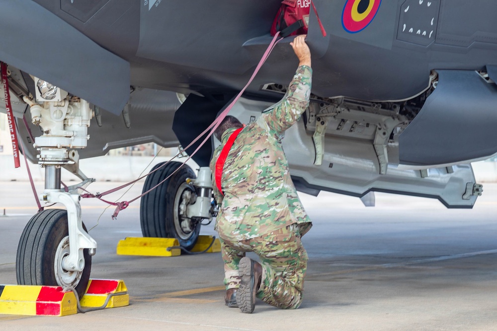 A military aircraft maintainer in camouflage uniform is kneeling beneath a gray stealth fighter jet, performing pre-flight or post-flight checks. The technician is handling red safety streamers/remove-before-flight tags on the underside of the aircraft. Yellow wheel chocks are visible securing the landing gear, indicating the aircraft is parked on a flight line.