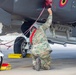 A military aircraft maintainer in camouflage uniform is kneeling beneath a gray stealth fighter jet, performing pre-flight or post-flight checks. The technician is handling red safety streamers/remove-before-flight tags on the underside of the aircraft. Yellow wheel chocks are visible securing the landing gear, indicating the aircraft is parked on a flight line.