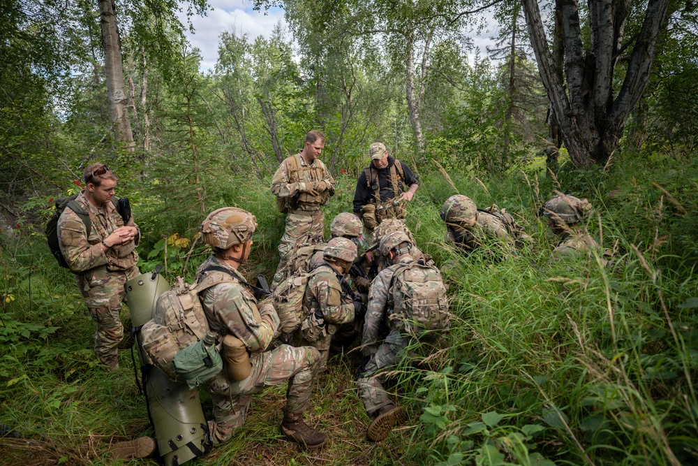 A group of approximately eight to nine military personnel in camouflage uniforms are gathered in a forested area, appearing to conduct a field training exercise or briefing. Most are wearing MultiCam/OCP pattern uniforms and tactical gear including helmets, body armor, and backpacks. One individual in dark civilian-style clothing appears to be instructing or observing the group.