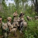 A group of approximately eight to nine military personnel in camouflage uniforms are gathered in a forested area, appearing to conduct a field training exercise or briefing. Most are wearing MultiCam/OCP pattern uniforms and tactical gear including helmets, body armor, and backpacks. One individual in dark civilian-style clothing appears to be instructing or observing the group.