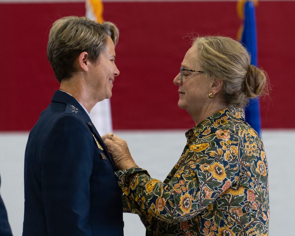 A formal military pinning ceremony is taking place, where an older civilian woman in a floral dress is pinning an insignia or medal onto the uniform of a female military officer in a dark blue dress uniform. Both women are smiling and facing each other in what appears to be an indoor venue with red and blue flag backdrops. The setting and attire suggest a promotion or award ceremony.