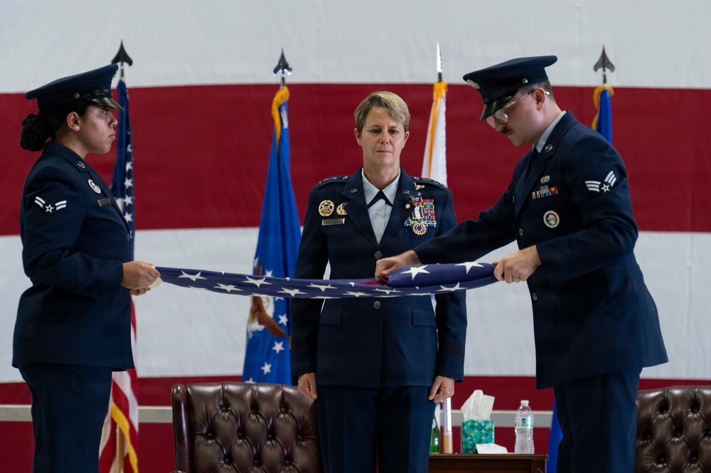 Three U.S. Air Force personnel are participating in a formal military flag-folding ceremony. A senior officer with multiple decorations and medals stands in the center while two lower-ranked airmen fold an American flag. The ceremony takes place in front of a large American flag backdrop with Air Force unit flags visible in the background.