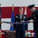 Three U.S. Air Force personnel are participating in a formal military flag-folding ceremony. A senior officer with multiple decorations and medals stands in the center while two lower-ranked airmen fold an American flag. The ceremony takes place in front of a large American flag backdrop with Air Force unit flags visible in the background.