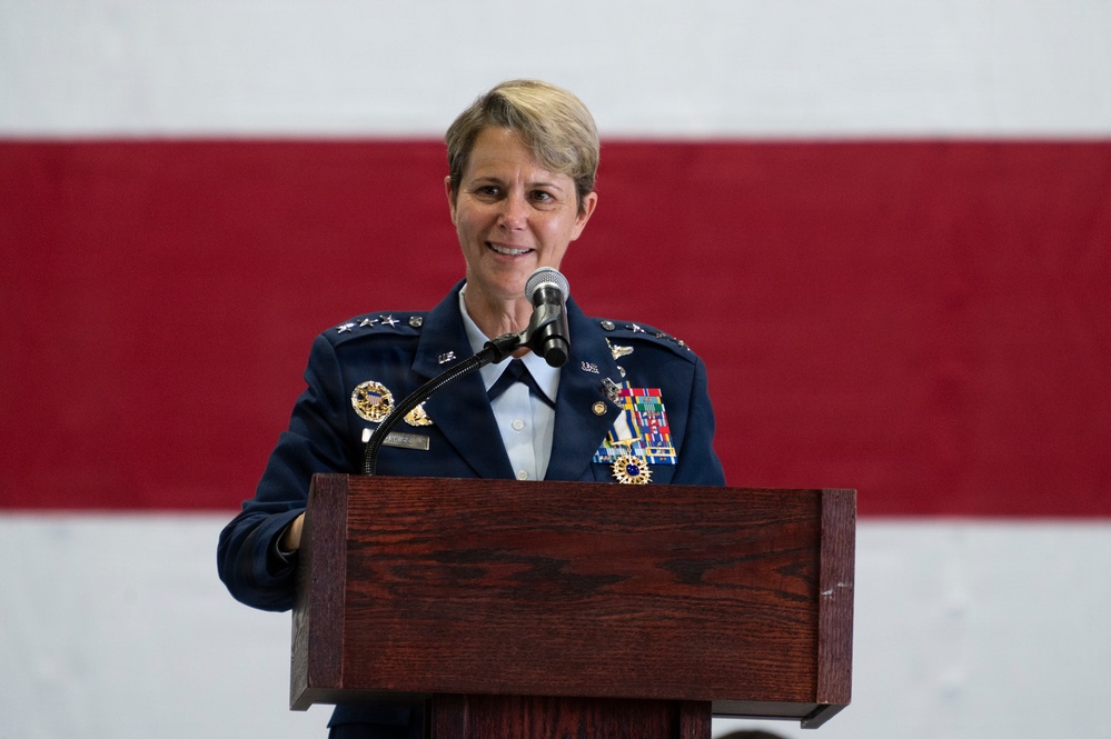 A female U.S. Air Force officer in dress blues is speaking at a wooden podium with a microphone. She is wearing a high-ranking uniform adorned with multiple medals, ribbons, and insignia, with three stars visible on her shoulder indicating the rank of Lieutenant General. An American flag is displayed prominently in the background.
