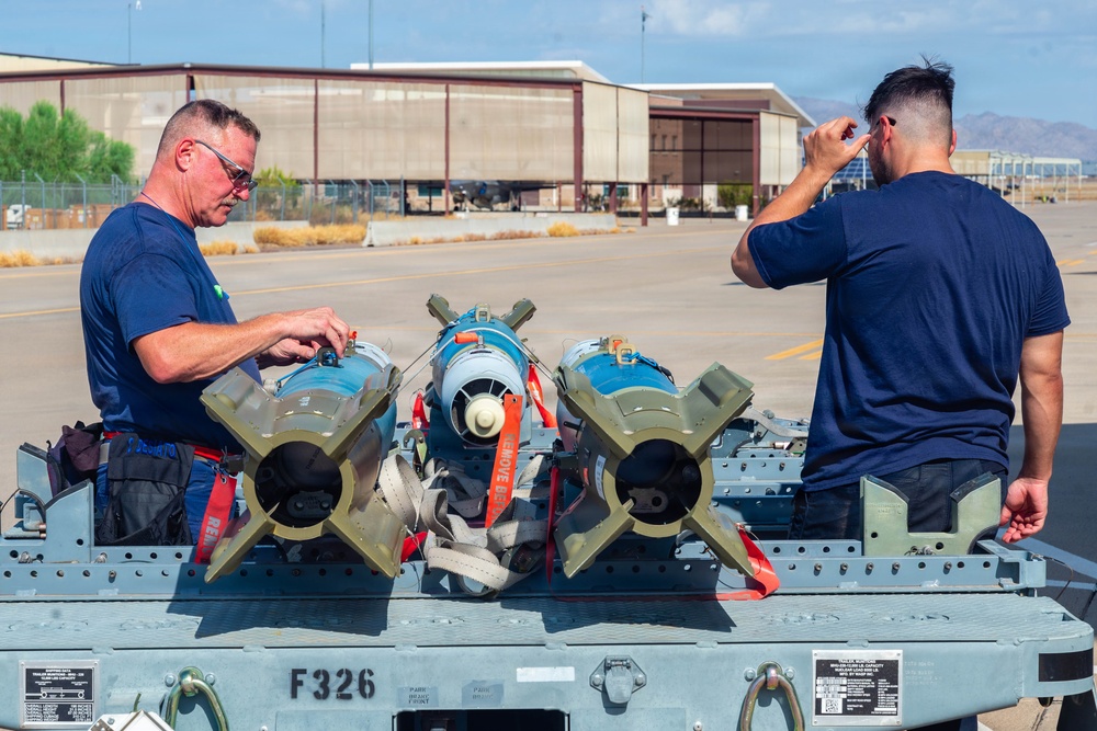 Two military personnel in navy blue t-shirts are working with munitions on a weapons cart labeled 'F326' on a military airfield flight line. Three blue-painted practice/training bombs with fin assemblies and red 'REMOVE BEFORE FLIGHT' safety streamers are secured on the cart. Large aircraft hangars and mountains are visible in the background, consistent with a desert military installation.