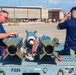 Two military personnel in navy blue t-shirts are working with munitions on a weapons cart labeled 'F326' on a military airfield flight line. Three blue-painted practice/training bombs with fin assemblies and red 'REMOVE BEFORE FLIGHT' safety streamers are secured on the cart. Large aircraft hangars and mountains are visible in the background, consistent with a desert military installation.