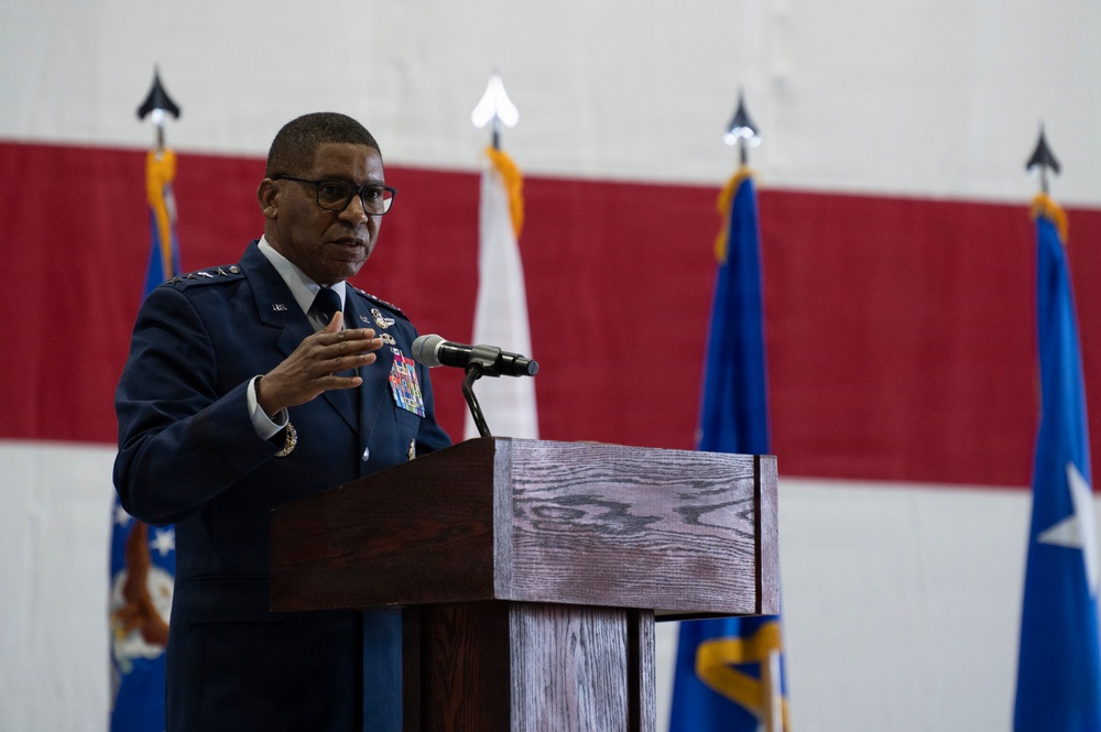 A high-ranking U.S. Air Force officer in dress blues is delivering a speech at a podium equipped with a microphone. The setting appears to be a military hangar or ceremonial hall, decorated with large American flag bunting and multiple Air Force flags. The officer's uniform displays numerous medals and insignia consistent with senior general officer rank.