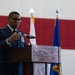 A high-ranking U.S. Air Force officer in dress blues is delivering a speech at a podium equipped with a microphone. The setting appears to be a military hangar or ceremonial hall, decorated with large American flag bunting and multiple Air Force flags. The officer's uniform displays numerous medals and insignia consistent with senior general officer rank.