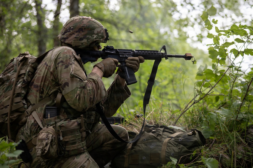A soldier in full combat gear is positioned in a kneeling stance in a wooded area, aiming an assault rifle forward. The soldier is wearing MultiCam (OCP) camouflage pattern uniform and helmet, consistent with U.S. Army equipment. The setting appears to be a training exercise or field operation in a dense forest environment.