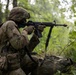 A soldier in full combat gear is positioned in a kneeling stance in a wooded area, aiming an assault rifle forward. The soldier is wearing MultiCam (OCP) camouflage pattern uniform and helmet, consistent with U.S. Army equipment. The setting appears to be a training exercise or field operation in a dense forest environment.