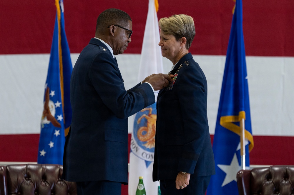A military ceremony in which a senior officer is pinning a medal or decoration onto another officer's uniform. Both individuals are wearing dark blue U.S. Air Force dress uniforms, and the event appears to be a formal awards or change-of-command ceremony. The backdrop includes a large American flag, Air Force flags, and what appears to be a command seal.