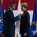 A military ceremony in which a senior officer is pinning a medal or decoration onto another officer's uniform. Both individuals are wearing dark blue U.S. Air Force dress uniforms, and the event appears to be a formal awards or change-of-command ceremony. The backdrop includes a large American flag, Air Force flags, and what appears to be a command seal.