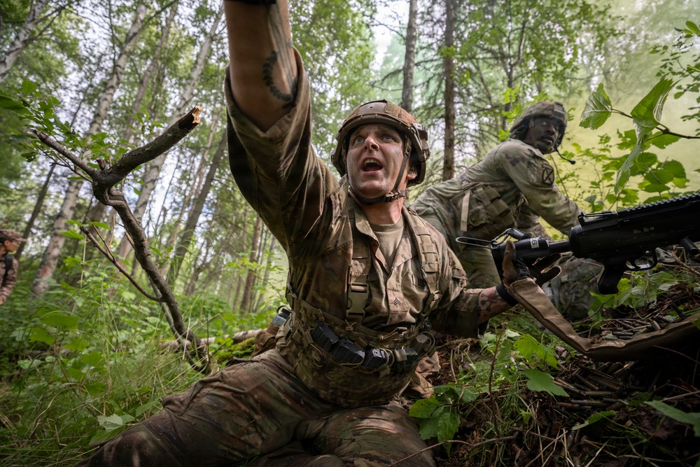 A U.S. Army soldier in multicam/OCP camouflage uniform is captured in a dynamic low-angle photograph in a forested training environment. The foreground soldier is gesturing with one raised arm while holding a rifle, appearing to be issuing commands or signals to fellow soldiers. The scene depicts what appears to be a military field training exercise in a dense woodland setting.