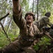 A U.S. Army soldier in multicam/OCP camouflage uniform is captured in a dynamic low-angle photograph in a forested training environment. The foreground soldier is gesturing with one raised arm while holding a rifle, appearing to be issuing commands or signals to fellow soldiers. The scene depicts what appears to be a military field training exercise in a dense woodland setting.