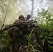 Two soldiers in full combat gear are taking cover behind a fallen tree in a dense forest environment. Colored smoke (appearing greenish-yellow) fills the background, suggesting a training exercise or simulated combat scenario. The foreground soldier appears to be shouting or giving commands while both personnel maintain low tactical positions.