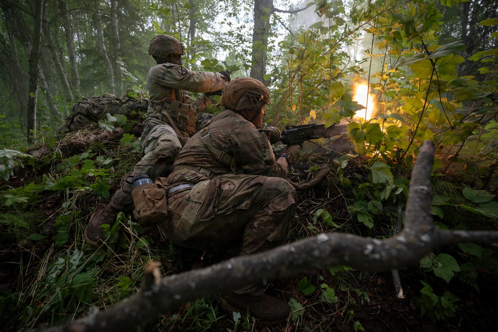 Two soldiers in full camouflage combat gear are positioned behind natural cover in a dense forest, with one actively firing a weapon producing a visible muzzle flash. The scene appears to be a military training exercise or combat operation conducted in a wooded environment. Both soldiers are wearing modern tactical equipment including helmets, body armor, and load-bearing vests.