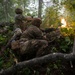 Two soldiers in full camouflage combat gear are positioned behind natural cover in a dense forest, with one actively firing a weapon producing a visible muzzle flash. The scene appears to be a military training exercise or combat operation conducted in a wooded environment. Both soldiers are wearing modern tactical equipment including helmets, body armor, and load-bearing vests.