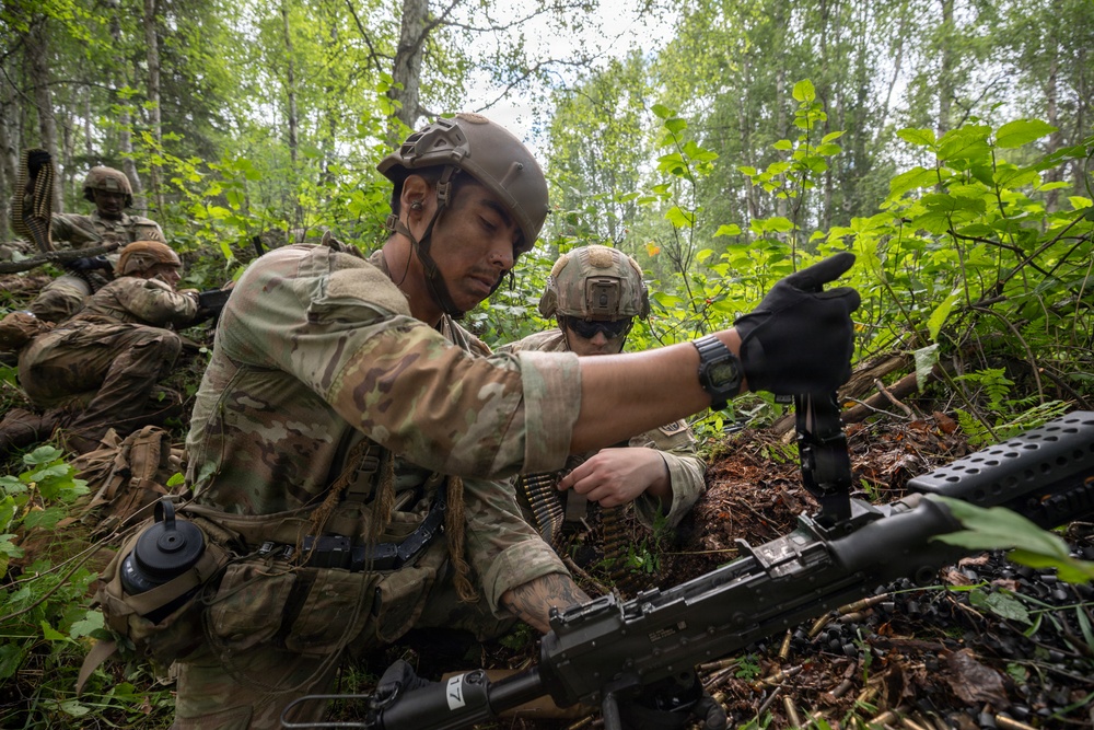 Military personnel in full combat gear are positioned in a forested environment, working with a belt-fed machine gun. The foreground soldier appears to be loading or handling an ammunition belt while another soldier points in a direction, suggesting a tactical training exercise or field operation. At least three soldiers are visible among dense woodland vegetation.