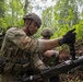 Military personnel in full combat gear are positioned in a forested environment, working with a belt-fed machine gun. The foreground soldier appears to be loading or handling an ammunition belt while another soldier points in a direction, suggesting a tactical training exercise or field operation. At least three soldiers are visible among dense woodland vegetation.