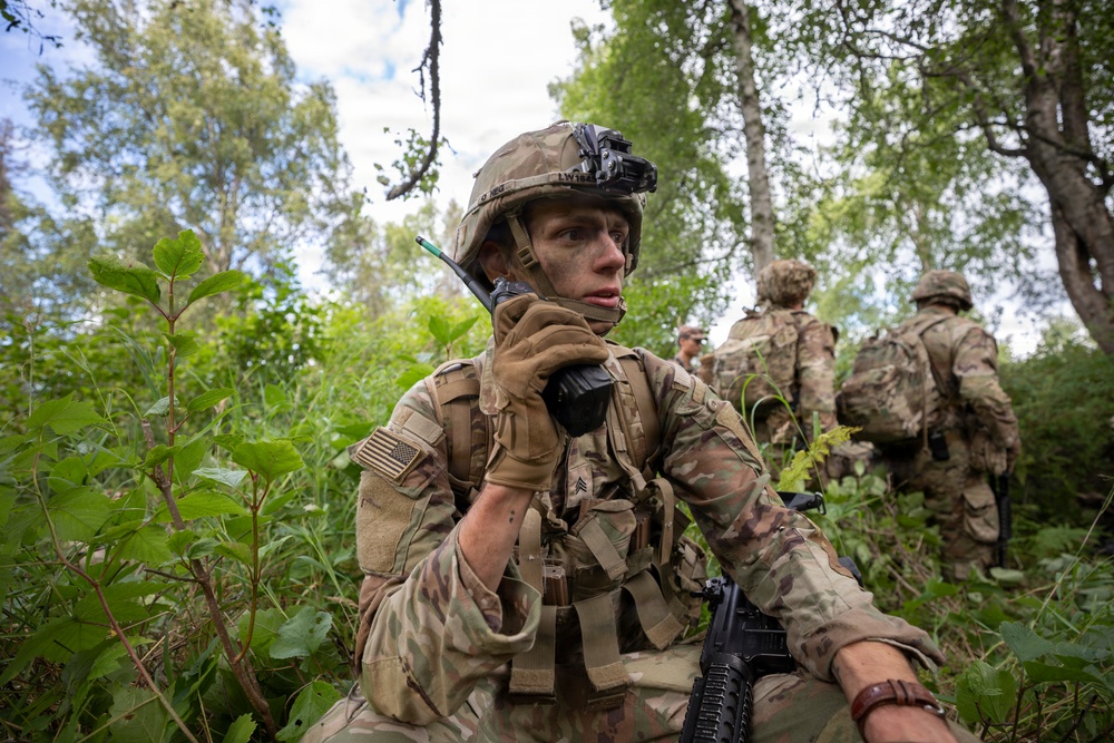 A U.S. Army sergeant in full combat gear is using a radio handset while positioned in a forested environment, with face camouflage paint applied. Two additional soldiers are visible in the background moving through the same wooded terrain. The scene appears to depict a field training exercise or tactical patrol operation.