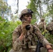 A U.S. Army sergeant in full combat gear is using a radio handset while positioned in a forested environment, with face camouflage paint applied. Two additional soldiers are visible in the background moving through the same wooded terrain. The scene appears to depict a field training exercise or tactical patrol operation.