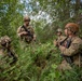 Four military personnel in full combat gear are gathered in a dense woodland area, appearing to conduct a briefing or tactical discussion. They are wearing OCP (Operational Camouflage Pattern) uniforms with helmets and carrying rifles and communication equipment. The setting suggests a field training exercise or operational patrol in a forested environment.