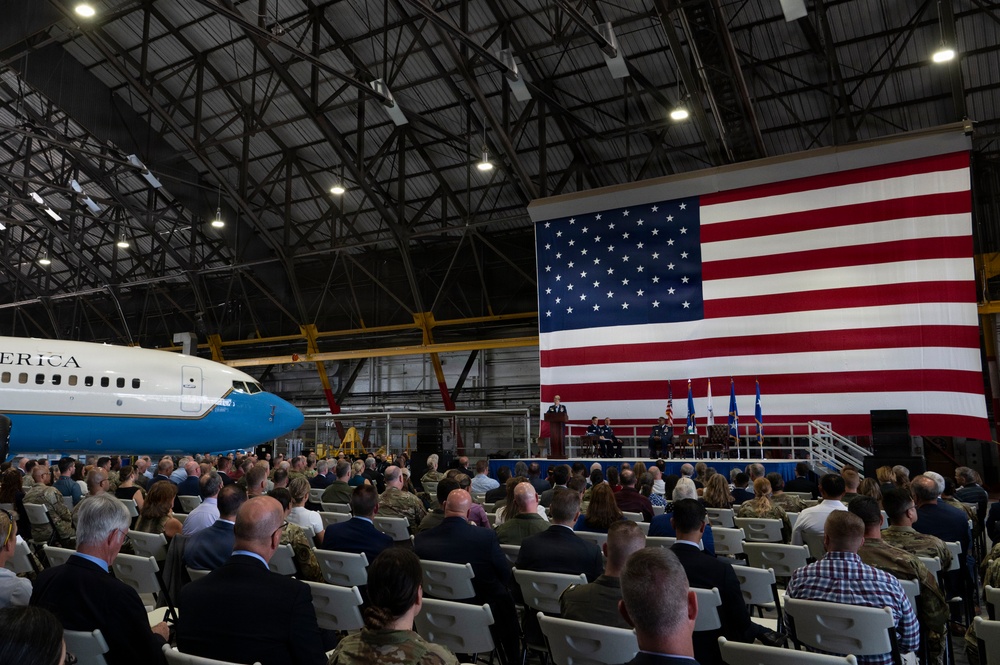 A formal military ceremony is taking place inside a large aircraft hangar. A massive American flag serves as a backdrop on the stage where uniformed military personnel are gathered at a podium, while a large audience of both military and civilian attendees is seated in white folding chairs. A distinctive blue and white aircraft with 'ERICA' visible on the fuselage — consistent with Air Force One livery — is parked to the left.