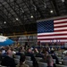 A formal military ceremony is taking place inside a large aircraft hangar. A massive American flag serves as a backdrop on the stage where uniformed military personnel are gathered at a podium, while a large audience of both military and civilian attendees is seated in white folding chairs. A distinctive blue and white aircraft with 'ERICA' visible on the fuselage — consistent with Air Force One livery — is parked to the left.
