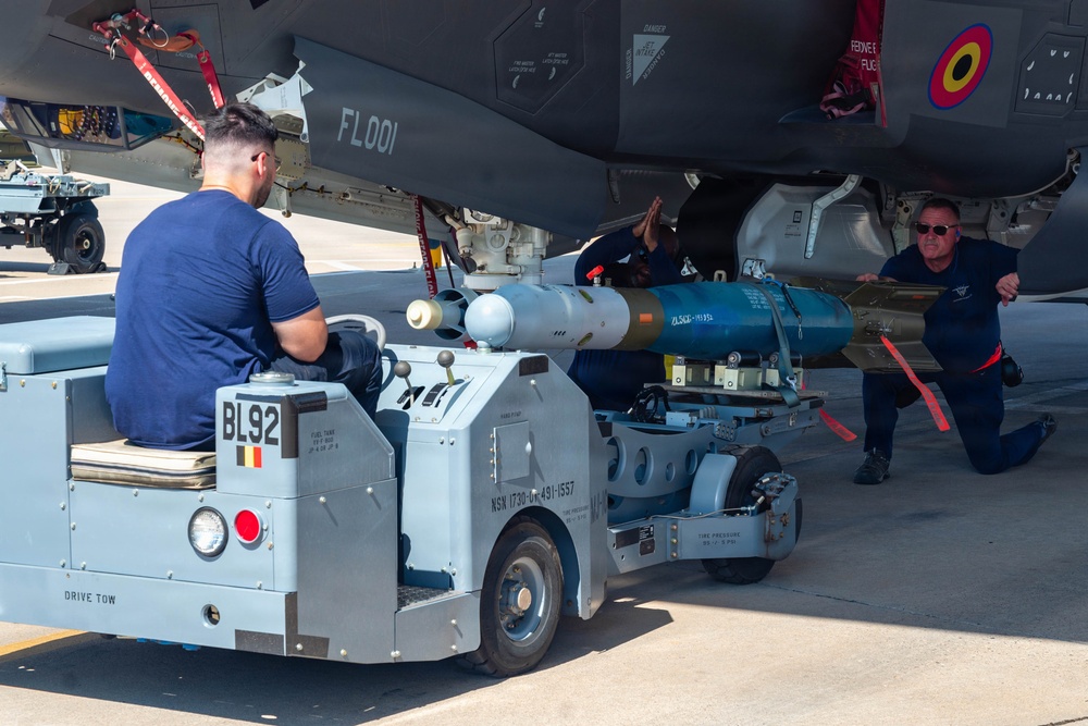 Ground crew personnel are loading or positioning munitions onto a military aircraft, identified by tail code 'FL001' and bearing a Belgian flag roundel. A weapons loading cart labeled 'BL92' is being used to lift and align what appears to be a guided bomb or missile under the aircraft's fuselage. At least three technicians are involved in the operation on what appears to be a flight line or tarmac.