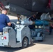 Ground crew personnel are loading or positioning munitions onto a military aircraft, identified by tail code 'FL001' and bearing a Belgian flag roundel. A weapons loading cart labeled 'BL92' is being used to lift and align what appears to be a guided bomb or missile under the aircraft's fuselage. At least three technicians are involved in the operation on what appears to be a flight line or tarmac.