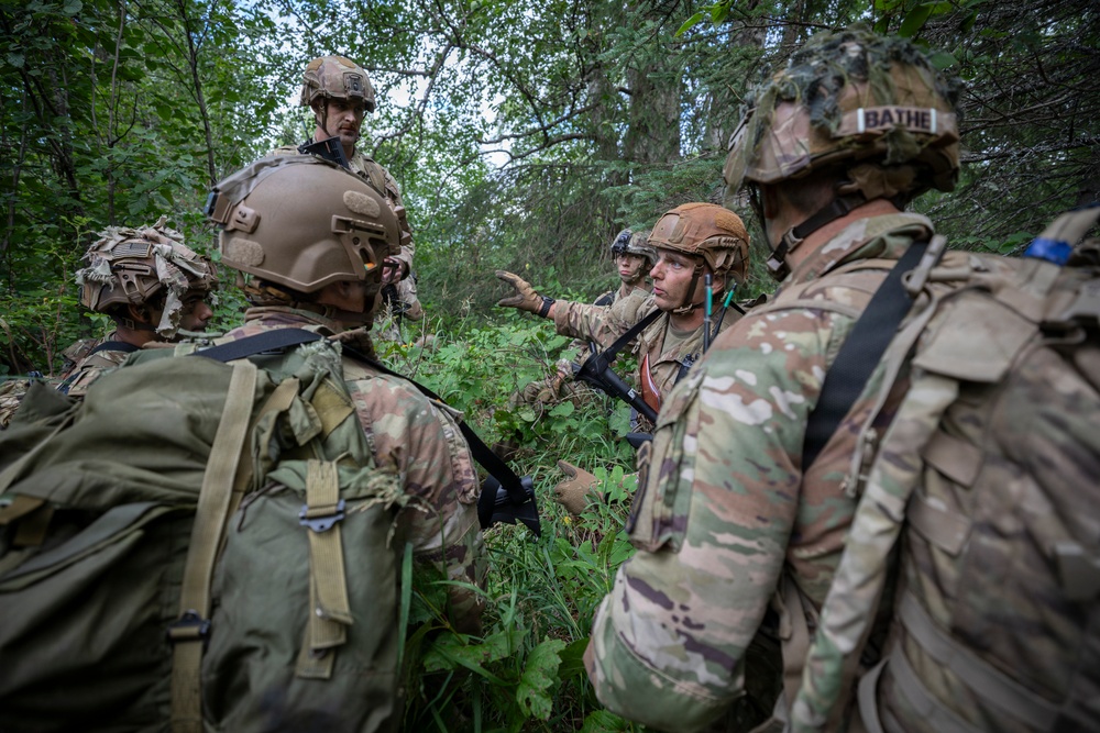A group of military personnel in full combat gear are gathered in a wooded area, appearing to conduct a tactical briefing or after-action review. One soldier in the center is gesturing and appears to be speaking to the group. The soldiers are wearing MultiCam/OCP camouflage uniforms with tactical helmets and rucksacks.