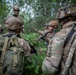 A group of military personnel in full combat gear are gathered in a wooded area, appearing to conduct a tactical briefing or after-action review. One soldier in the center is gesturing and appears to be speaking to the group. The soldiers are wearing MultiCam/OCP camouflage uniforms with tactical helmets and rucksacks.
