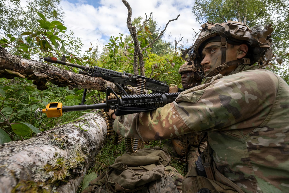 Two soldiers in MultiCam camouflage uniforms are positioned behind a fallen birch tree in a wooded area, operating what appears to be an M249 or M240 belt-fed machine gun. The foreground soldier has camouflage face paint applied and is aiming the weapon over the log, with a visible ammunition belt loaded into the weapon. A yellow training/blank-firing adapter is attached to the muzzle, indicating this is a military training exercise rather than live combat.
