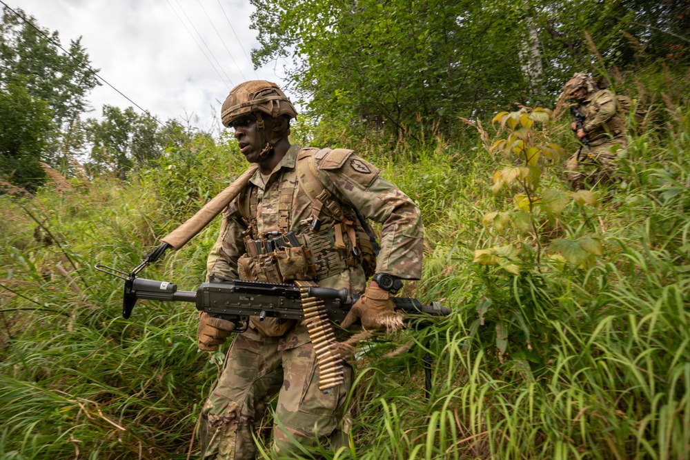 Two soldiers in full combat gear are moving through dense vegetation and tall grass in what appears to be a military training exercise or field operation. The lead soldier is prominently carrying a light machine gun with an ammunition belt, wearing OCP (Operational Camouflage Pattern) uniform and tactical vest. A second soldier is visible in the background, also armed and in similar gear.
