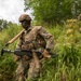Two soldiers in full combat gear are moving through dense vegetation and tall grass in what appears to be a military training exercise or field operation. The lead soldier is prominently carrying a light machine gun with an ammunition belt, wearing OCP (Operational Camouflage Pattern) uniform and tactical vest. A second soldier is visible in the background, also armed and in similar gear.