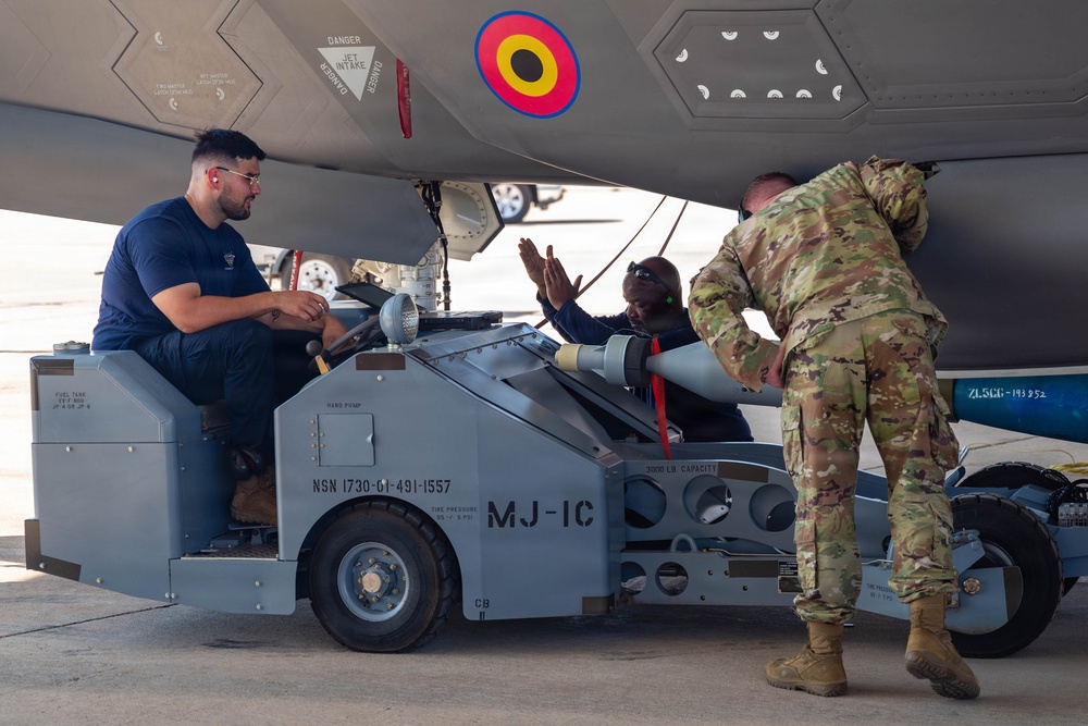 Military personnel are loading or handling a munition/missile beneath the fuselage of a fighter aircraft using an MJ-1C weapons loader. Three individuals are visible — one in civilian/navy blue attire operating the loader, and two in military camouflage uniforms assisting with the ordnance. A roundel in red, yellow, and blue colors is visible on the aircraft's underside, suggesting a foreign or allied air force aircraft.
