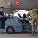 Military personnel are loading or handling a munition/missile beneath the fuselage of a fighter aircraft using an MJ-1C weapons loader. Three individuals are visible — one in civilian/navy blue attire operating the loader, and two in military camouflage uniforms assisting with the ordnance. A roundel in red, yellow, and blue colors is visible on the aircraft's underside, suggesting a foreign or allied air force aircraft.