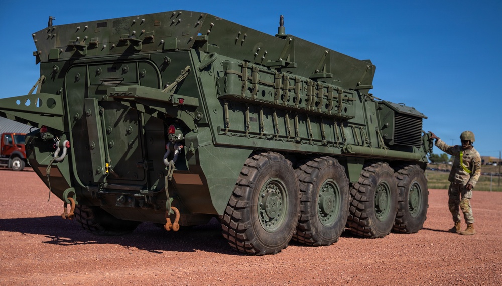 A large olive-drab military armored vehicle, consistent with a Stryker-family or similar 8-wheeled armored personnel carrier, is parked on a red dirt surface under a clear blue sky. A soldier in camouflage uniform and helmet is standing beside the vehicle on the right side, appearing to interact with or inspect it. A second individual is partially visible through an open side hatch of the vehicle.