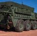 A large olive-drab military armored vehicle, consistent with a Stryker-family or similar 8-wheeled armored personnel carrier, is parked on a red dirt surface under a clear blue sky. A soldier in camouflage uniform and helmet is standing beside the vehicle on the right side, appearing to interact with or inspect it. A second individual is partially visible through an open side hatch of the vehicle.