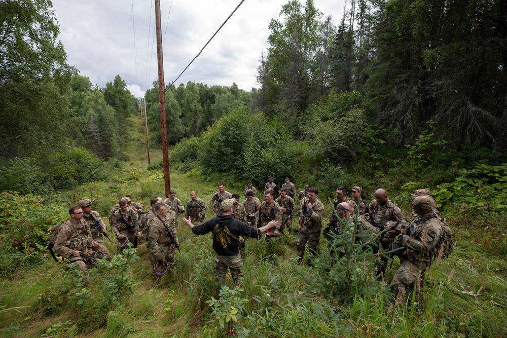 A group of approximately 20 military personnel in camouflage uniforms are gathered in a forested clearing beneath a utility power line corridor. A single individual in a dark civilian-style jacket stands with arms outstretched, appearing to brief or instruct the assembled soldiers. The setting appears to be a summer training exercise in a northern or boreal forest environment.