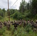 A group of approximately 20 military personnel in camouflage uniforms are gathered in a forested clearing beneath a utility power line corridor. A single individual in a dark civilian-style jacket stands with arms outstretched, appearing to brief or instruct the assembled soldiers. The setting appears to be a summer training exercise in a northern or boreal forest environment.