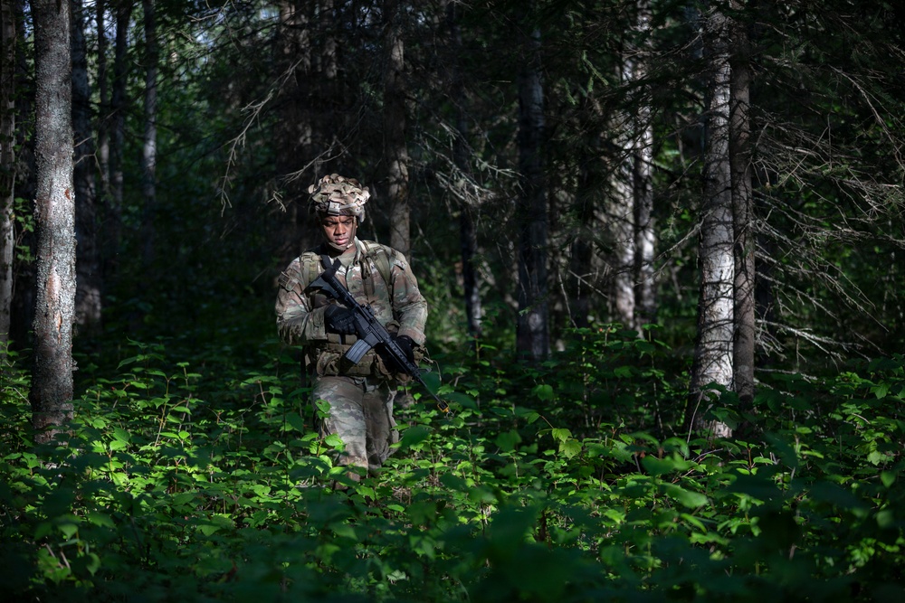 A soldier in full camouflage gear patrols through a dense forest with thick green undergrowth. The individual is equipped with a combat helmet, tactical vest, and carries an assault rifle. The setting appears to be a woodland training or operational environment during daylight hours.