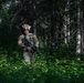 A soldier in full camouflage gear patrols through a dense forest with thick green undergrowth. The individual is equipped with a combat helmet, tactical vest, and carries an assault rifle. The setting appears to be a woodland training or operational environment during daylight hours.