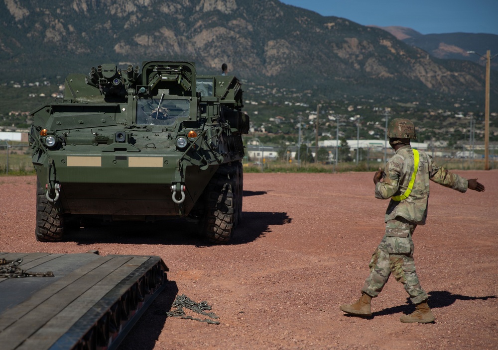 A U.S. Army soldier wearing a high-visibility safety vest and combat helmet is directing a Stryker armored fighting vehicle on a gravel surface, likely at a military installation. The vehicle appears to be maneuvering near a flatbed rail car or transport platform, suggesting a loading or unloading operation. A mountainous landscape consistent with the Colorado Front Range is visible in the background.