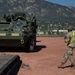 A U.S. Army soldier wearing a high-visibility safety vest and combat helmet is directing a Stryker armored fighting vehicle on a gravel surface, likely at a military installation. The vehicle appears to be maneuvering near a flatbed rail car or transport platform, suggesting a loading or unloading operation. A mountainous landscape consistent with the Colorado Front Range is visible in the background.