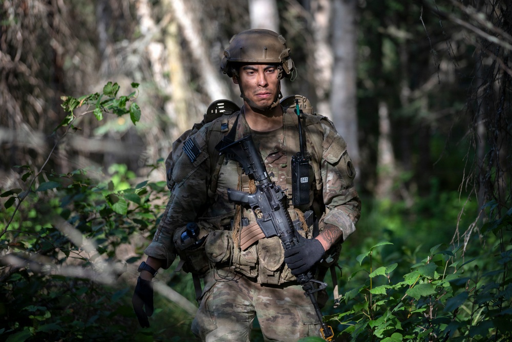 A soldier in full combat gear moves through a forested environment, carrying an assault rifle and wearing multicam camouflage uniform with tactical vest. The individual appears to be on patrol or participating in a field training exercise, navigating dense woodland terrain. The image is professionally composed with sharp focus on the subject against a softly blurred forest background.