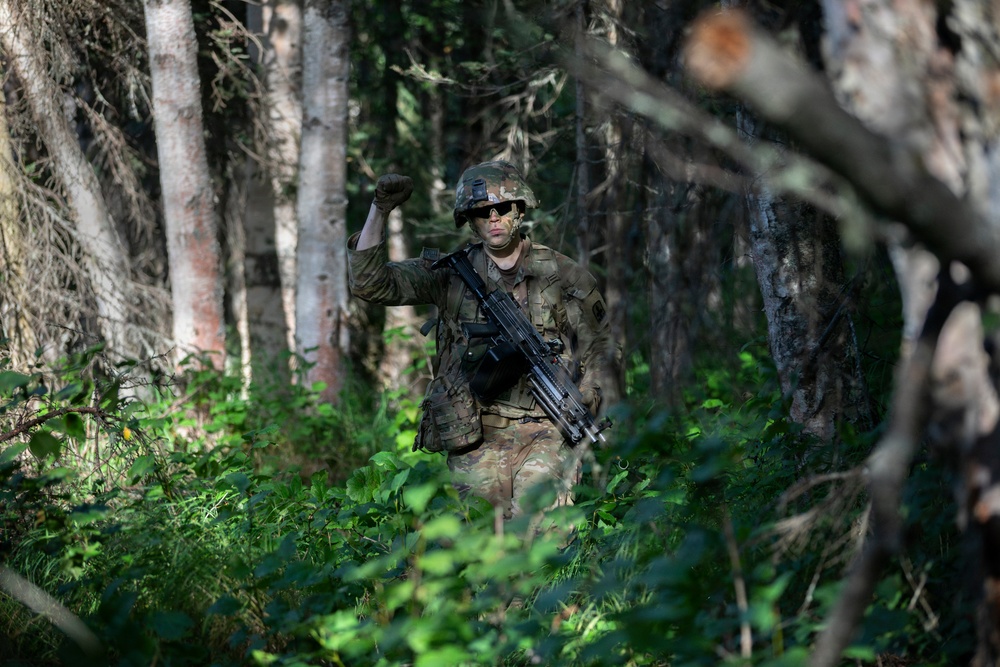 A soldier in full combat gear moves through a dense forest environment. The soldier is giving a hand signal with a raised fist, indicating a halt or stop signal to other personnel. The individual is equipped with camouflage uniform, tactical helmet, face paint, and carries an assault rifle.