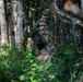 A soldier in full combat gear moves through a dense forest environment. The soldier is giving a hand signal with a raised fist, indicating a halt or stop signal to other personnel. The individual is equipped with camouflage uniform, tactical helmet, face paint, and carries an assault rifle.