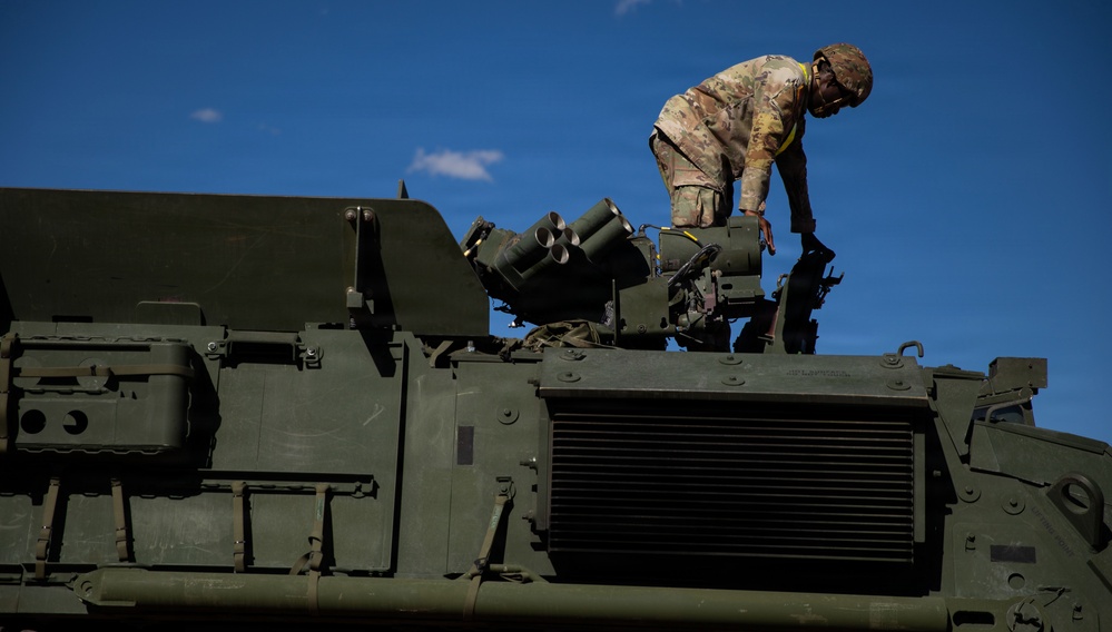 A soldier in camouflage uniform is working on top of a large military armored vehicle, likely performing maintenance or equipment checks. The vehicle is olive drab green and features visible weapon systems including what appear to be grenade launchers or smoke dischargers. The image is taken from a low angle against a clear blue sky.